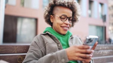 Kahkahalar atan, yeşil kapüşonlu, akıllı telefonuyla yazan ve dışarıdaki kameraya bakan Afrikalı kadın.
