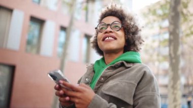 Yeşil kapüşonlu olumlu Afrikalı kadın etrafa bakıyor ve dışarıda akıllı telefon tutuyor.