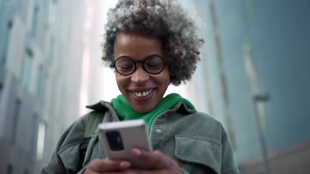 笑顔大人アフリカの女性は屋外の建物の近くの電話でカジュアルな服を着てテキストメッセージ — ストック動画