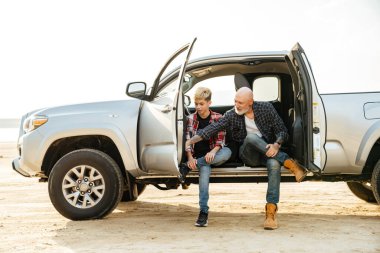 Gülümseyen baba ve oğlu sahilde arabada oturmuş konuşuyor.