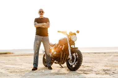 Dışarıda motosikletle poz verirken kameraya bakan, dövmeli, olgun, cesur bir adam.