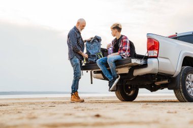 Beyaz, orta yaşlı bir baba ve anaokulu çocuğu plajda park etmiş bir arabanın arkasında kamp malzemesi tutuyorlar.