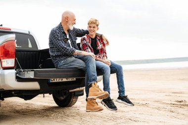 Gülümseyen baba ve oğlu sahilde bir arabanın bagajında oturuyor.