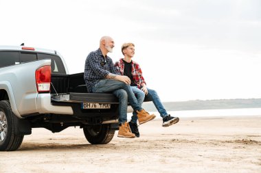 Gülümseyen baba ve oğlu sahilde bir arabanın bagajında oturuyor.