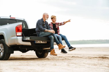 Gülümseyen baba ve oğlu bir arabanın bagajında oturuyor kumsalın arkasında.