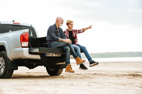 Gülümseyen baba ve oğlu bir arabanın bagajında oturuyor kumsalın arkasında.