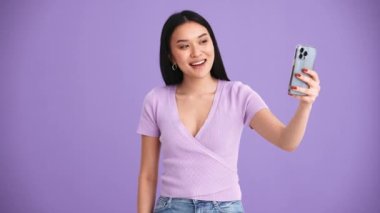 Mor tişört giyen gülümseyen Asyalı esmer kadın mor stüdyoda cep telefonuyla selfie çekiyor.