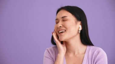 Gülümseyen Asyalı esmer kadın mor tişört giyip mor stüdyoda kulaklıkla müzik dinliyor.