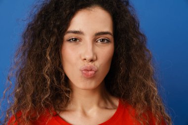 Genç esmer kadın öpüşürken dudaklarını öpüyor ve mavi duvardan izole edilmiş kameraya bakıyor.