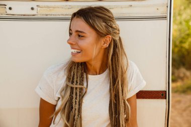 Açık havada karavana yaslanırken saçları örgülü beyaz bir kadın gülümsüyor.