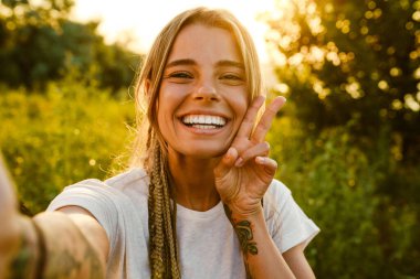 Beyaz bir kadın dışarıda fotoğraf çekerken gülümsüyor ve el kol hareketi yapıyor.