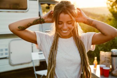 At kuyruklu beyaz bir kadın dışarıda karavanın yanında dururken gülümsüyor.