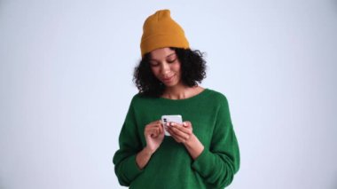 Gri stüdyoda şapka takan kıvırcık saçlı bir kadın telefon aracılığıyla telefon ediyor.