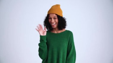 Gülümseyen kıvırcık saçlı kadın gri stüdyoda selam veriyor.