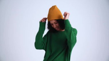 Gri stüdyoda şapkalı, kıvırcık saçlı, yakışıklı bir kadın dans ediyor.