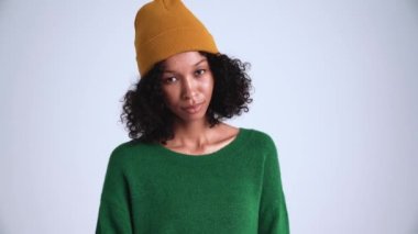 Yakışıklı, kıvırcık saçlı, şapkalı bir kadın gri stüdyoda kameraya bakıyor.