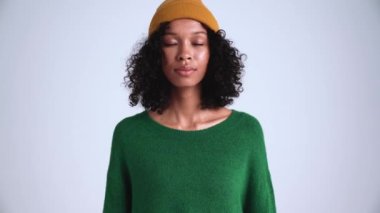 Yoğunlaşmış kıvırcık saçlı, şapkalı kadın gri stüdyodaki kameraya bakıyor.