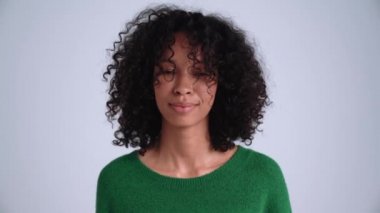 Gülümseyen kıvırcık saçlı kadın gri stüdyodaki kameraya sessizlik işareti yapıyor.