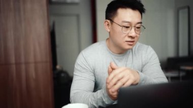 Gözlük takan Asyalı özgüvenli adam evdeki bilgisayardan video ile konuşuyor.