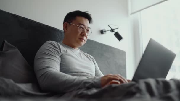 Sonriente Hombre Asiático Con Gafas Trabajo Ordenador Portátil Cama Casa — Vídeos de Stock