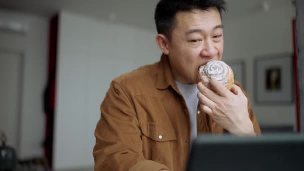 Lächelnder Asiatischer Mann Isst Brötchen Und Schaut Hause Video Auf — Stockvideo