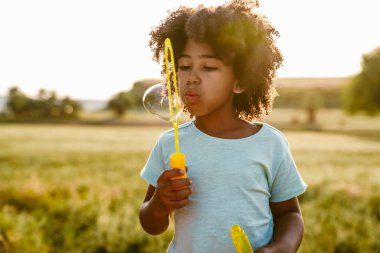 Siyah kıvırcık tişörtlü çocuk dışarıda sabun köpüğü yaparken gülümsüyor.