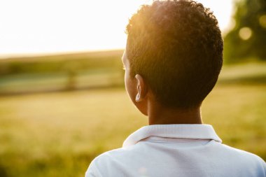 Beyaz tişörtlü siyah çocuk yaz tarlasında dururken gün batımını seyrediyor.