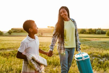 Siyah mutlu aile yaz tarlasında çantalarla yürürken gülümsüyor.