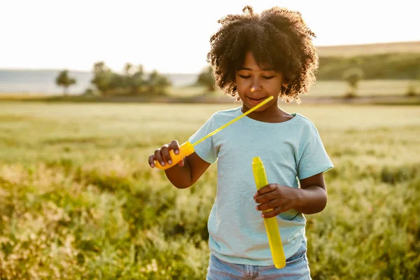 Siyah kıvırcık tişörtlü çocuk dışarıda sabun köpüğü yaparken gülümsüyor.