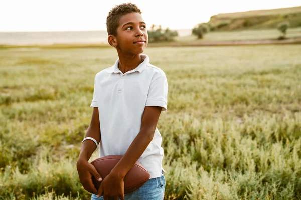 Beyaz tişörtlü siyah çocuk dışarıda rugby topuyla poz veriyor.