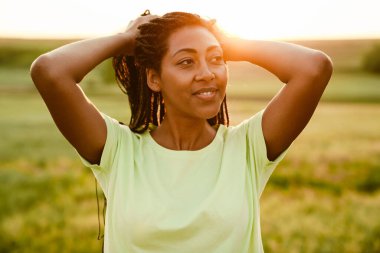 At kuyruklu siyahi kadın, dışarıda dikilirken gülümsüyor ve yan bakıyor.