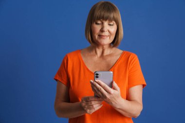 Mavi duvarda izole edilmiş cep telefonu kullanan üst düzey sarışın kadın tişörtü giyiyor.