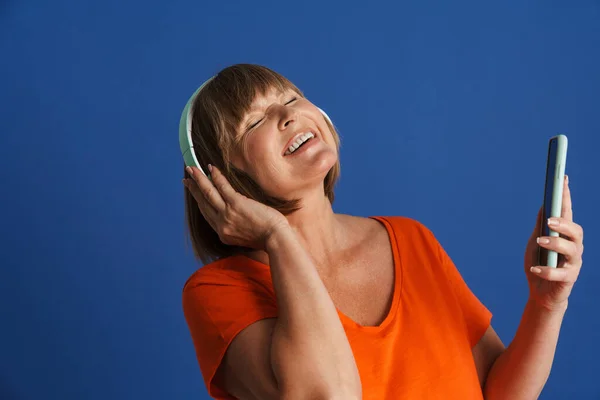 年长的金发女人一边听着音乐一边笑着 耳机被隔离在蓝色的墙上 — 图库照片