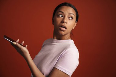 Kırmızı duvarda izole edilmiş cep telefonu kullanan genç siyah bir kadın tişört giyiyor.