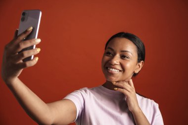 Kırmızı duvardan izole edilmiş cep telefonuyla selfie çekerken gülümseyen genç siyahi kadın.