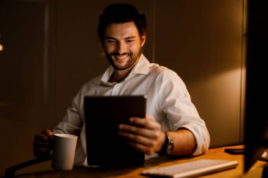 Ofiste tablet bilgisayarla çalışırken kahve içen beyaz sakallı adam.