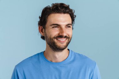 Tüylü genç esmer adam gülümsüyor ve mavi arka planda soyutlanmış bir şekilde yan tarafa bakıyor.