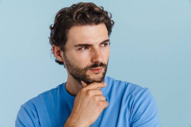 Genç esmer, kıllı bir adam mavi arka planda yalnız poz veriyor ve yan bakıyor.