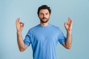 Mavi arka planda meditasyon yaparken tüylü, esmer genç bir adam.