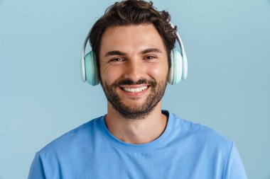 Kıllı saçlı genç adam mavi arka planda izole edilmiş kulaklıklarla müzik dinliyor.