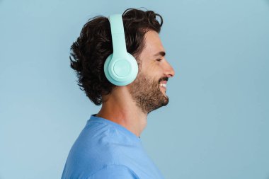Kıllı saçlı genç adam mavi arka planda izole edilmiş kulaklıklarla müzik dinliyor.