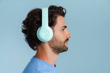 Kıllı saçlı genç adam mavi arka planda izole edilmiş kulaklıklarla müzik dinliyor.