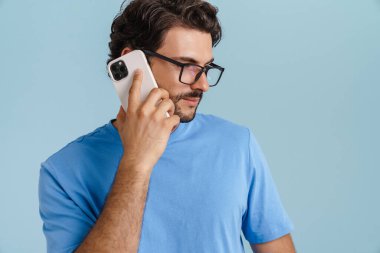 Gözlüklü genç esmer adam mavi arka planda cep telefonuyla konuşuyor.