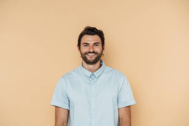 Genç beyaz adam gülümsüyor ve bej arka planda izole edilmiş kameraya bakıyor.