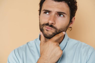 Gömlek giyen genç beyaz bir adam çenesini tutuyor ve bej arka planda kapalı bir kenara bakıyor.