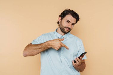 Beyaz kıllı adam bej arka planda izole edilmiş cep telefonuna işaret ediyor.