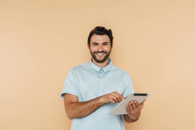 Beyaz kıllı adam gülümsüyor ve bej arkaplanda izole edilmiş tablet bilgisayar kullanıyor