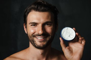 Siyah arka planda izole edilmiş yüz kremi sergilerken gülümseyen yarı çıplak genç bir adam.