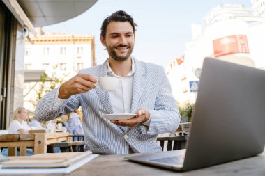 Genç adam dışarıda kafede kahve içerken dizüstü bilgisayarla çalışıyor.