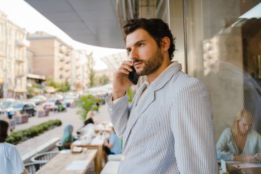 Dışarıda kafede dikilen genç adam cep telefonuyla konuşuyor.